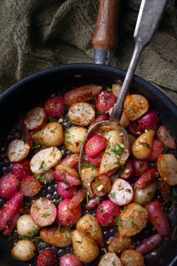 Fried radishes of different colors with herbs and spices on an iron frying pan with a spoon. Vegetarian food