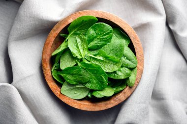 Baby spinach leaves with water drops in a wooden bowl on linen tablecloth. Raw organic spinach greens close up