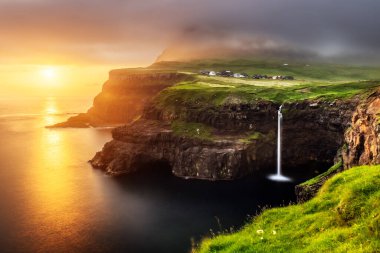Gün batımında Mulafossur şelalesi. Altın ışıkla çarpıcı bir gökyüzü. Gasadalur Köyü, Vagar Adası, Faroe Adaları, Danimarka. Peyzaj fotoğrafçılığı