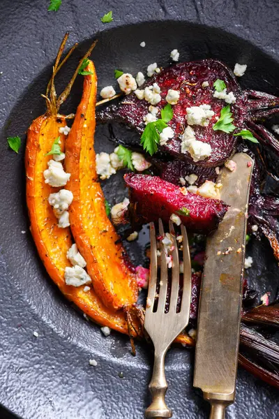 stock image Baked beet and carrot with goat cheese in black plate with knife and fork. Top view