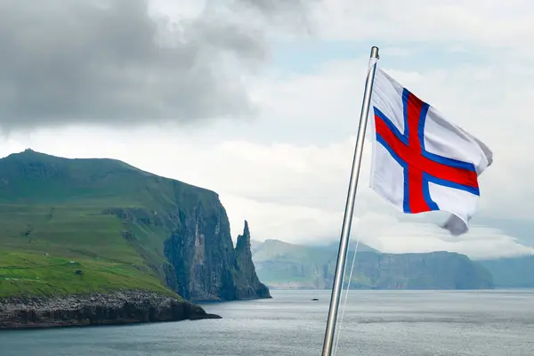 stock image National Faroese flag waving against a Witches Finger cliffs from Trollkonufingur viewpoint. Vagar island, Faroe Islands, Denmark. Landscape photography