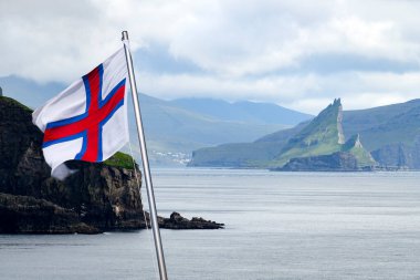 View of the Drangarnir and Tindholmur sea stacks from a boat sailing from the city of Sorvagur to the island of Mykines. Flag of Faroe islands waving against a cloudy sky. Landscape photography clipart