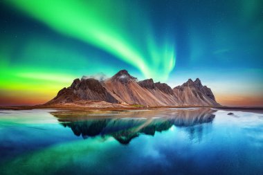 Aurora borealis kuzey ışıkları Vestrahorn Burnu 'ndaki ünlü Stokksnes dağlarının üzerinde. Gökyüzünün arka planındaki berrak suda yansıma, İzlanda. Peyzaj fotoğrafçılığı