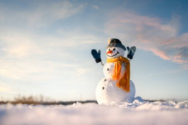 Örgü şapkalı gülen kardan adam ve elleri karlı tarlada olan sarı atkı. Arkaplanda bulutlarla ılık gün batımı gökyüzü