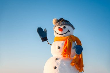 Örgü şapkalı gülen kardan adam ve elleri karlı tarlada olan sarı atkı. Arka planda mavi gökyüzü