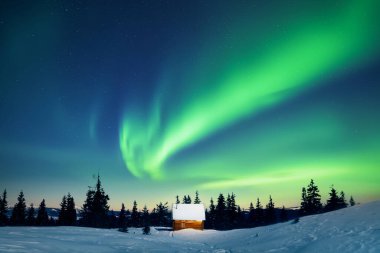 Karlı dağlardaki küçük ahşap evin üzerinde inanılmaz kuzey ışıkları. Noel tatili ve kış tatili konsepti. Aurora Borealis. Kutup ışıkları