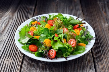 Kırsal ahşap arka planda beyaz bir tabakta taze yeşillik ve kiraz domateslerinin renkli salata karışımı. Yemek tasarımı ilham verici.
