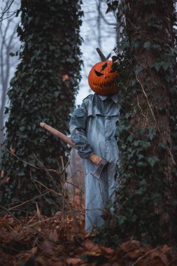 Sonbahar ormanlarında elinde balkabağı kafası ve balta silahıyla ürkünç korkuluk. Karanlık Cadılar Bayramı konsepti orman korkusu ve ürpertici gerilimle dolu.