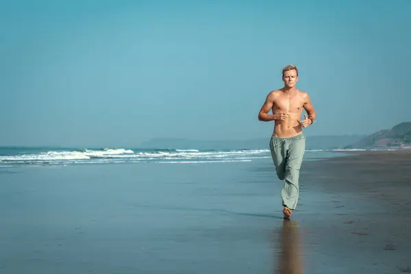 ビーチの濡れた砂に沿ってシャツレスで筋肉質の男がジョギングし 背景には海と山があります 彼は彼の顔に激しく集中した表情を持っている 海岸でジョギング ロイヤリティフリーのストック画像