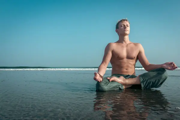 シャツのない若者がビーチの濡れた砂の上に瞑想的な蓮の上に座り 広い海の波が彼の後ろに墜落する ロイヤリティフリーのストック写真