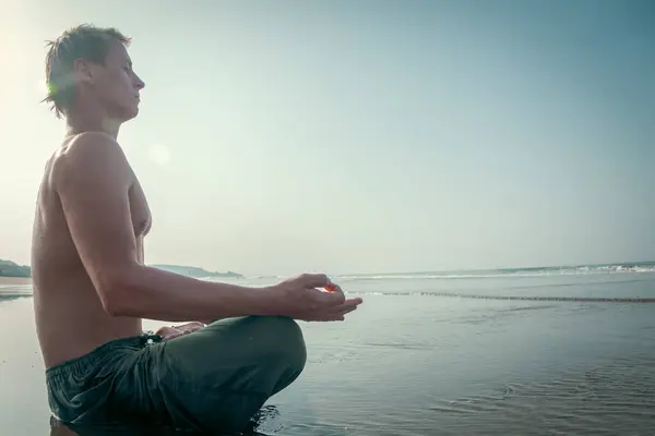 シャツのない若者がビーチの濡れた砂の上に瞑想的な蓮の上に座り 広い海の波が彼の後ろに墜落する ロイヤリティフリーのストック画像