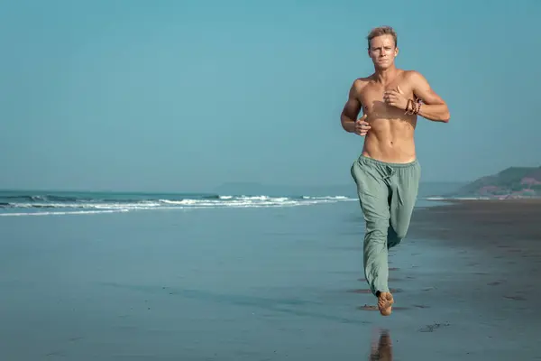 ビーチの濡れた砂に沿ってシャツレスで筋肉質の男がジョギングし 背景には海と山があります 彼は彼の顔に激しく集中した表情を持っている 海岸でジョギング ストック画像