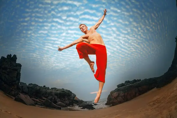 赤いズボンのシャツのない男は 自由と冒険の感覚を除いて 岩の崖と砂浜で 劇的な空に向かって喜んでジャンプします ロイヤリティフリーのストック画像