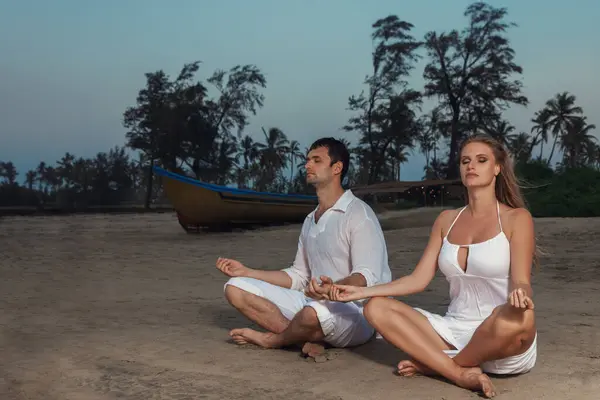 Young couple relaxing in lotus yoga position on sandy beach near colorful boat during sunset.