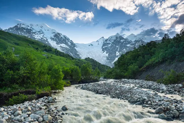 Renkli taşlarla dolu nehir akıntısı. Beyaz tepeleri ve ormanları olan dağlar. Yaz manzarası. Bulutlu mavi gökyüzü. Buzullu bir fotoğraf. Yukarı Svaneti, Gürcistan, Avrupa.