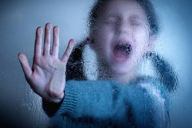 Conceptual image: Protect children and young underage people from violence, exploitation, abuse, and neglect. Close up young girl showing stop gesture from behind wet glass. Selective focus on hand.