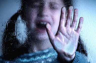 Dramatic portrait of scared young girl making NO sign, showing hand STOP gesture. Kid standing behind wet glass and says No to domestic violence, abuse, school bullying,  problem concept