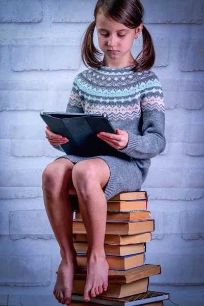 Young girl sitting on stack of books with PC tablet. Concept of learning new knowledge, self improvement and development of mental abilities. Vertical image.