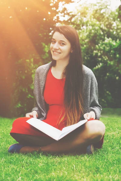 Beautiful woman reading book in nature. Young attractive student in red dress studying and reading a book.