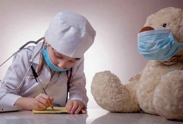 给病人泰迪熊开药方的戴着防护面罩的可爱小女孩医生的画像 保健和医药概念 — 图库照片
