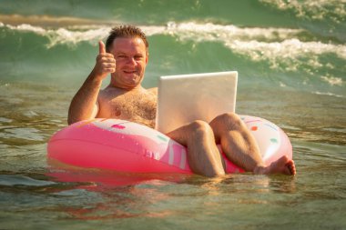 Yaz iş hayalleri. Okyanusta ya da denizde dizüstü bilgisayarla yüzen bir iş adamı. Yaz tatili. Uzaktan çalışan ve şişirilebilir bir tekerleğin üzerinde dinlenen mutlu iş adamının komik portresi.. 