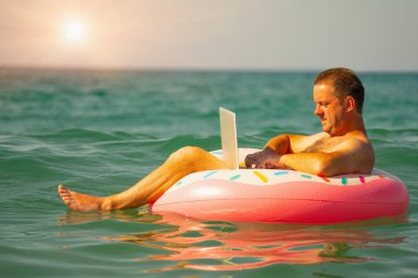 Uzaktan kumandayla ilgili rüyalar. Okyanusta ya da denizde dizüstü bilgisayarla yüzen işadamlarının yan görüntüsü. Mutlu iş adamının portresi kışın çalışıyor ve şişme bir tekerleğin üzerinde dinleniyor.. 