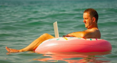 Uzaktan kumandayla ilgili rüyalar. Okyanusta ya da denizde dizüstü bilgisayarla yüzen işadamlarının yan görüntüsü. Mutlu iş adamının portresi kışın çalışıyor ve suda şişirilebilir bir tekerleğin üzerinde dinleniyor. Yatay resim.