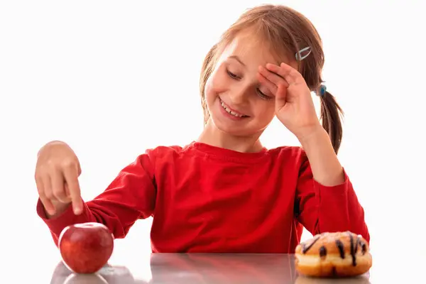 Pretty young girl choose between sweet cake and apple. Child pointing on apple with finger as if she want to say: healthy food it\'s really good. Horizontal image.