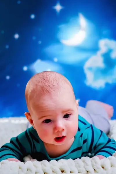Time to sleep, carefree dreams concept. Funny portrait of baby boy 4 months old lies on the bed against night sky background.