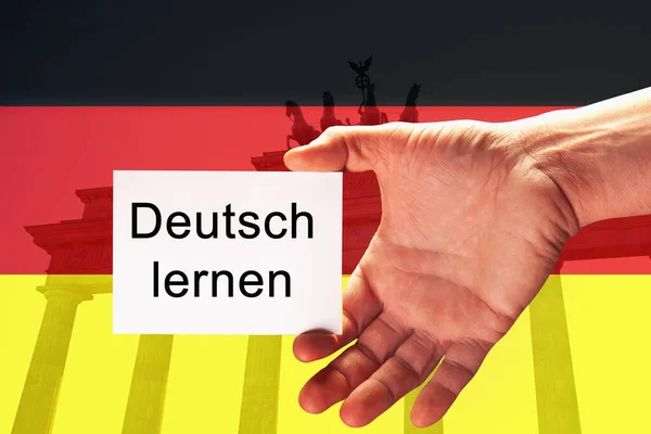 Closeup hand of young man holding white card with description: Deutsch lernen (LEARN GERMAN), written in german, against background of German flag and Brandenburg Gate in Berlin