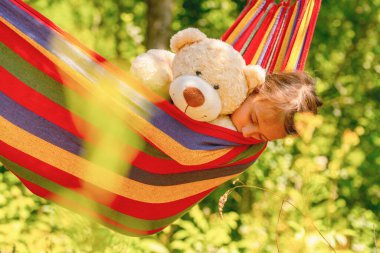 Yaz tatili ve dinlenme konsepti. Oyuncak ayıyla genç ve güzel bir kız yeşil bahçedeki hamakta uyuyor. Yatay resim.