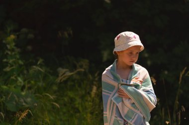 Deniz sahilinde, bitkilerin koyu yeşil arka planına karşı havluyla kaplanmış güzel bir kız. Tasarım için alanı kopyala