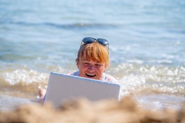 Work anywhere and anytime. Portrait of very happy business woman lying in the water and working online with laptop on the beach of sea or ocean. clipart