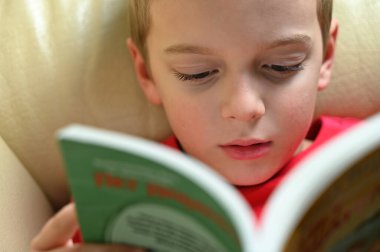 Genç çocuk evde dinleniyor, kanepede kitap okuyor.