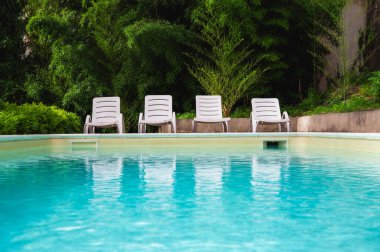 Merdiven ve mavi suyla yüzme havuzu. Mavi yüzme havuzunun yanındaki yeşil yemyeşil bahçedeki beyaz güneş yatakları. Yaz rahatlama konsepti. Su yüzeyinin dokusu. Metin için boşluk.