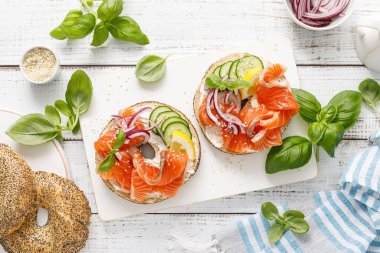 Salmon sandwiches with bagel, salted fish, fresh cucumber, onion and basil on white background, top view. Healthy breakfast with salmon toasts