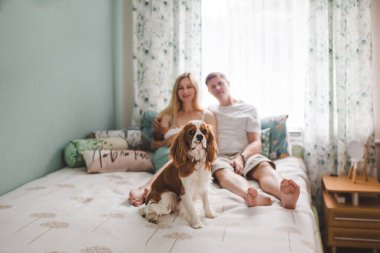 Genç bir aile yatak odasında rahat bir şekilde oturuyor, anne yeni doğmuş bebeklerini kucağında tutarken Kral Charles Spaniel ön planda oturuyor ve sahneye sıcaklık katıyor..