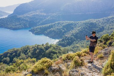 Kayalık yolda duran erkek turist, Türkiye 'nin Mula ilinin Fethiye ilçesinde mavi deniz ve yeşil tepelerle çarpıcı kıyı manzaralarının fotoğrafını çekiyor.