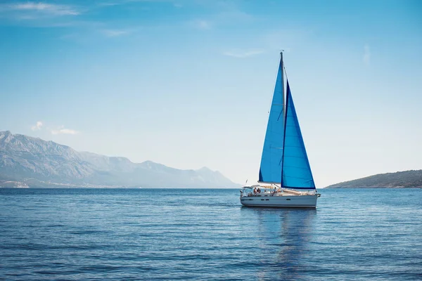 stock image White yacht with blue sails in the sea against a background of blue sky and mountains