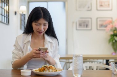 Bir restoranda, genç bir kadın telefonuyla mutlu bir fotoğraf çekiyor, modern yaşam tarzını benimsiyor.