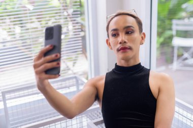 Bıyıklı, ruj sürmüş, tulumlu, buzlu kahve ve yeşillikli bir kafede cep telefonuyla selfie çeken güzel Asyalı tuhaf LGBT halk destekçisi portresi.