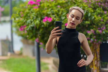 Güzel Asyalı ibne LGBT halk destekçisi bıyıklı, rujlu ve tulumlu parktaki açık havada telefon kullanan adam.