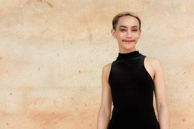 Güzel Asyalı tuhaf LGBT topluluk destekçisi ruj sürmüş ve tulum giymiş bej duvar arkasına gülümseyen adam portresi.