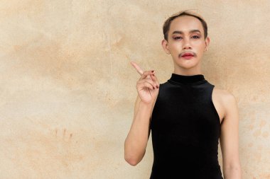 Güzel Asyalı tuhaf LGBT topluluk destekçisi ruj sürmüş ve tulum giyip bej duvarlara yaslanmış, reklam için telif alanını işaret eden adam portresi.
