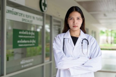 Hastane kliniğinin önünde beyaz laboratuvar önlüğü ve steteskop giymiş Asyalı genç kadın doktor.