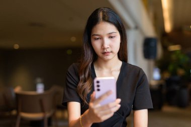 Genç Asyalı bir kadın kahve dükkanında oturuyor, şık, siyah bir elbise giyiyor ve cep telefonu kullanıyor.