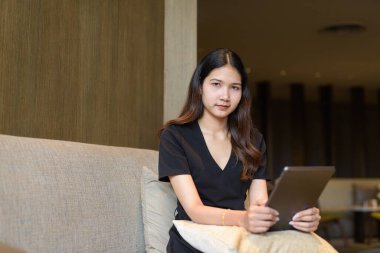 Genç Asyalı kadın kahve dükkanında oturuyor. Dijital tablet kullanarak şık, siyah bir elbise giyiyor.