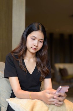 Genç Asyalı bir kadın kahve dükkanında oturuyor. Şık, siyah bir elbise giyiyor. Telefon kullanıyor.