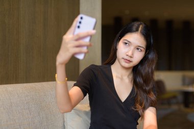 Genç Asyalı bir kadın kahve dükkanında oturuyor. Şık, siyah bir elbise giyiyor. Telefon kullanıyor.