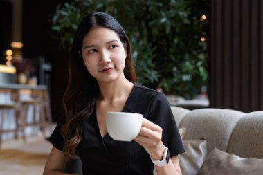 Genç Asyalı iş kadını kahve dükkanında oturmuş şık siyah elbiseler giyiyor ve kahve içiyor.